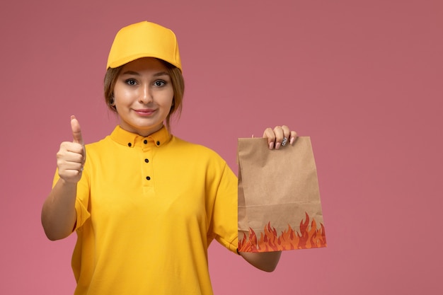 Mensajero femenino de vista frontal en capa amarilla uniforme amarillo sosteniendo el paquete de alimentos y mostrando como signo sobre fondo rosa trabajo de entrega uniforme trabajo de color
