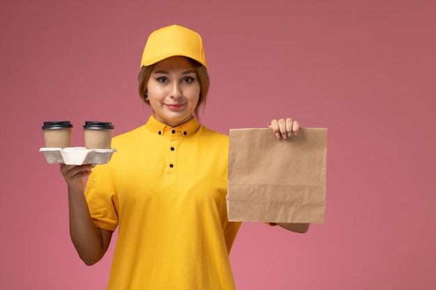 Mensajero femenino de vista frontal en capa amarilla uniforme amarillo sosteniendo el paquete de alimentos y tazas de café en color de trabajo femenino de entrega uniforme de escritorio rosa