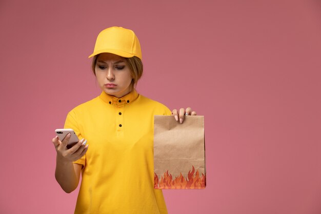 Mensajero femenino de vista frontal en capa amarilla uniforme amarillo sosteniendo el paquete de alimentos usando un teléfono sobre fondo rosa trabajo de entrega uniforme trabajo de color