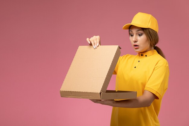 Mensajero femenino de vista frontal en capa amarilla uniforme amarillo sosteniendo el paquete de entrega abriéndolo en el trabajo de entrega uniforme de fondo rosa