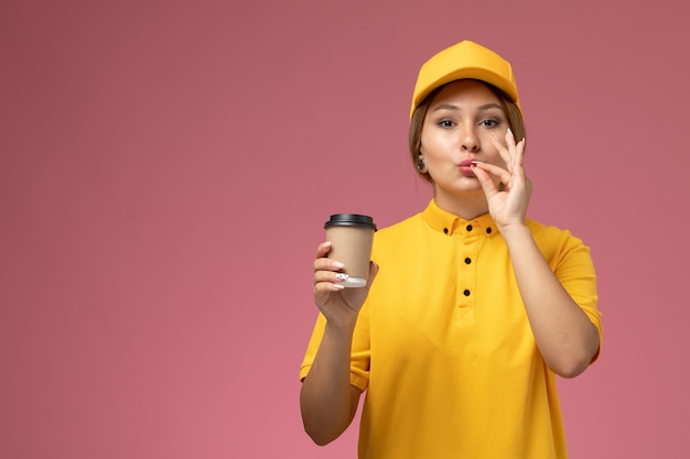 Mensajero femenino de vista frontal en capa amarilla uniforme amarillo sosteniendo una taza de café marrón de plástico que muestra signo sabroso en el color femenino de entrega uniforme de escritorio rosa