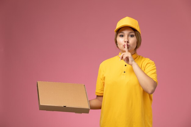 Mensajero femenino de vista frontal en capa amarilla uniforme amarillo sosteniendo una taza de café marrón de plástico que muestra el signo de silencio en el color femenino de entrega uniforme de escritorio rosa