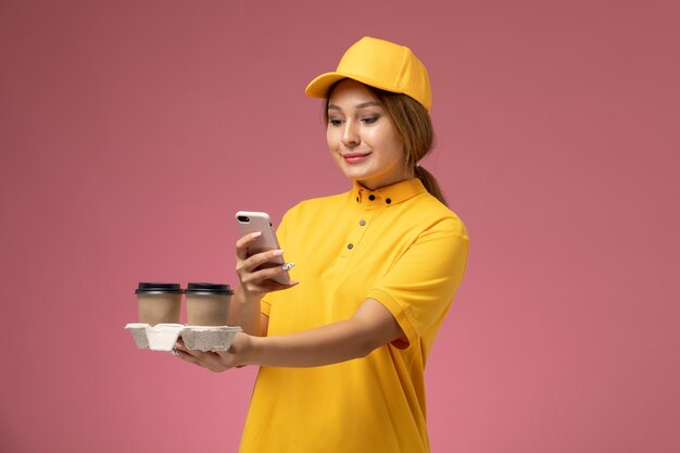Mensajero femenino de vista frontal en capa amarilla uniforme amarillo sosteniendo tazas de café con teléfono sobre fondo rosa trabajo de entrega uniforme trabajo de color