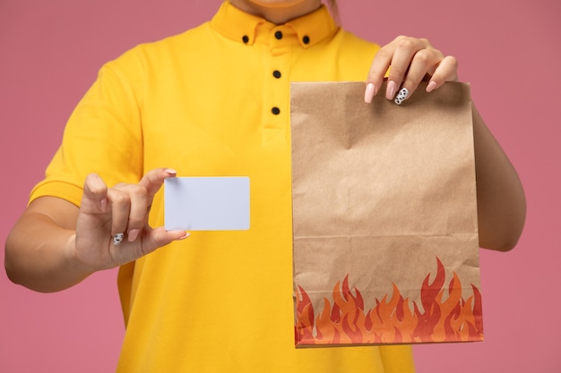 Mensajero femenino de vista frontal en capa amarilla uniforme amarillo con tarjeta blanca y paquete de alimentos en el trabajo de color de trabajo de entrega uniforme de escritorio rosa