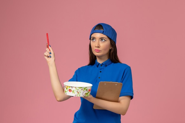 Mensajero femenino de vista frontal en capa uniforme azul con bloc de notas y bolígrafo con tazón de entrega en la pared rosa claro, servicio de entrega de empleados