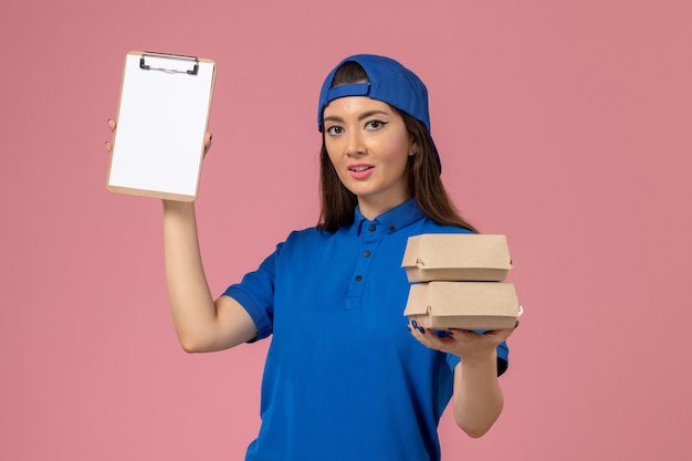 Mensajero femenino de vista frontal con capa uniforme azul con bloc de notas y pequeños paquetes de entrega en la pared rosa claro, entrega de empleados de servicio