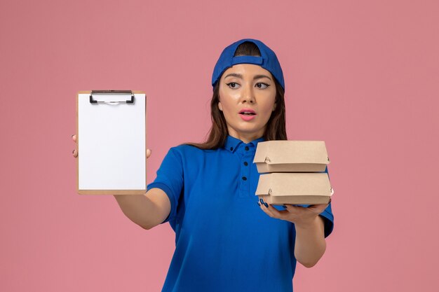 Mensajero femenino de vista frontal con capa uniforme azul con bloc de notas y pequeños paquetes de entrega en la pared rosa claro, entrega de empleados de servicio