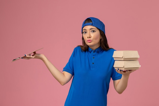 Mensajero femenino de vista frontal con capa uniforme azul con bloc de notas y pequeños paquetes de entrega en la pared rosa claro, entrega de empleados de servicio