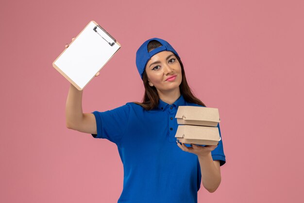 Mensajero femenino de vista frontal con capa uniforme azul con bloc de notas y pequeños paquetes de entrega en la pared rosa claro, entrega de empleados