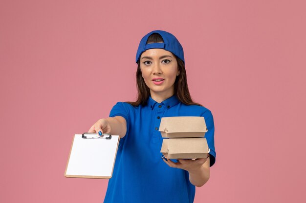 Mensajero femenino de vista frontal en capa uniforme azul con bloc de notas y pequeños paquetes de entrega en la pared rosa claro, trabajo de entrega de empleados de servicio