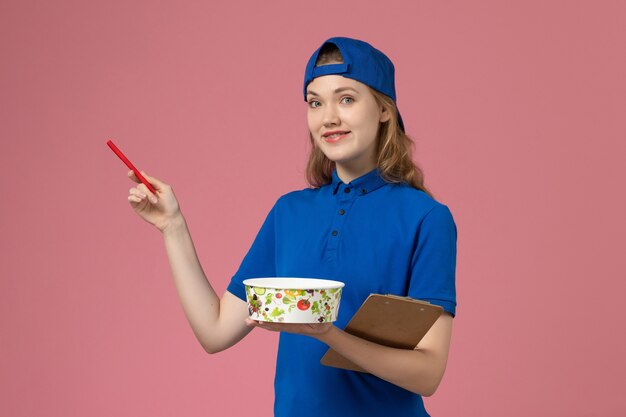Mensajero femenino de vista frontal en capa uniforme azul con bol de entrega y bolígrafo en la pared rosa claro, empleado de servicio de trabajo