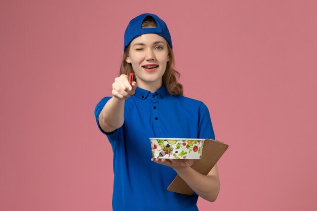 Mensajero femenino de vista frontal en capa uniforme azul con cuenco de entrega y bolígrafo de bloc de notas en la pared rosa claro, empleado de trabajo de entrega de servicio
