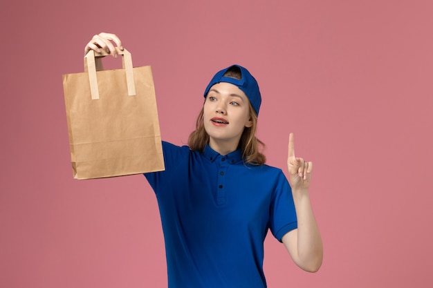 Mensajero femenino de vista frontal en capa uniforme azul con paquete de entrega de papel en la pared rosa, prestación de servicios para empleados