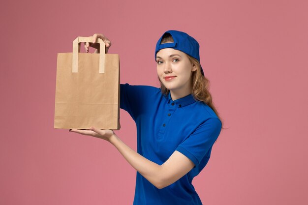 Mensajero femenino de vista frontal en capa uniforme azul con paquete de entrega de papel en la pared rosa, trabajador empleado de entrega de servicios