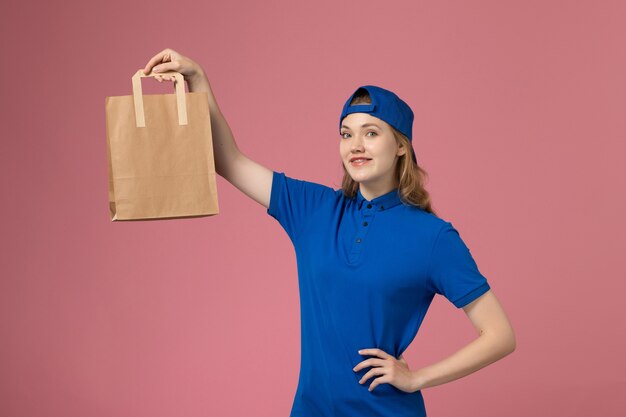 Mensajero femenino de vista frontal en capa uniforme azul con paquete de entrega de papel en la pared rosa, trabajo de empleado de entrega de servicio