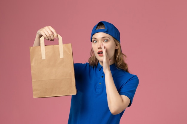 Mensajero femenino de vista frontal en capa uniforme azul con paquete de entrega de papel susurrando en la pared rosa, empleado de entrega de servicio