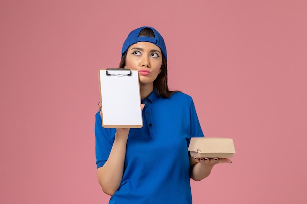 Mensajero femenino de vista frontal en capa uniforme azul con paquete de entrega pequeño vacío con bloc de notas pensando en la pared rosa, entrega de la empresa de servicios para empleados