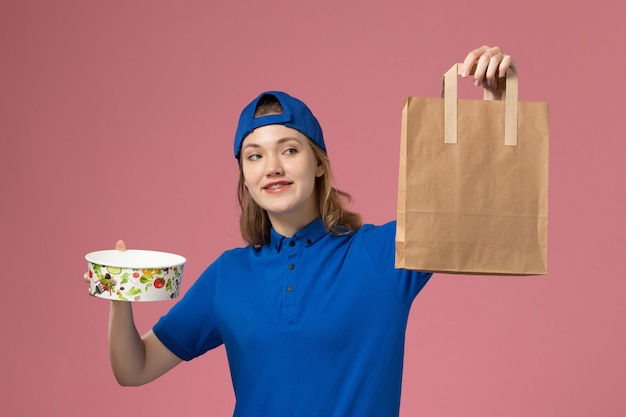 Mensajero femenino de vista frontal en capa uniforme azul con paquete de entrega y tazón en la pared rosa, trabajo de entrega de servicios para empleados