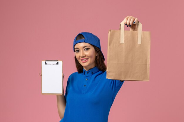 Mensajero femenino de vista frontal con capa uniforme azul con paquete de papel de entrega y bloc de notas en la pared rosa, empleado de trabajo de servicio entregando