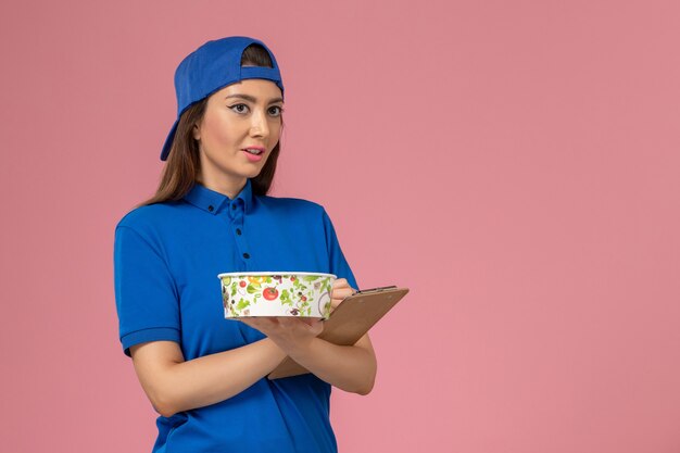 Mensajero femenino de vista frontal en capa uniforme azul sosteniendo el bloc de notas con el cuenco de entrega escribiendo notas en la pared rosa claro, servicio de entrega de empleados