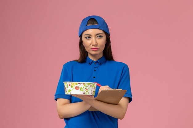 Mensajero femenino de vista frontal en capa uniforme azul sosteniendo el bloc de notas con el cuenco de entrega escribiendo notas en la pared rosa claro, trabajo de entrega del empleado de servicio