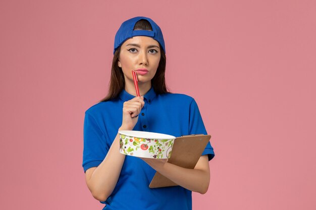 Mensajero femenino de vista frontal en capa uniforme azul sosteniendo el bloc de notas con cuenco de entrega pensando en la pared de color rosa claro, entrega de empleados de trabajo de servicio