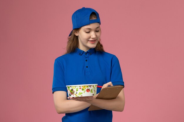 Mensajero femenino de vista frontal en capa uniforme azul sosteniendo el cuenco de entrega y el bloc de notas escribiendo en la pared rosa claro, empleado de entrega de servicio