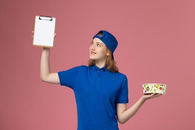 Mensajero femenino de vista frontal en capa uniforme azul sosteniendo el cuenco de entrega y el bloc de notas en la pared rosa claro, trabajador empleado de entrega de servicios