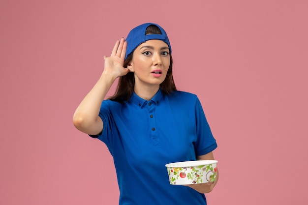 Mensajero femenino de vista frontal en capa uniforme azul sosteniendo el cuenco de entrega en la pared rosa claro, entrega de empleados de servicio de trabajo