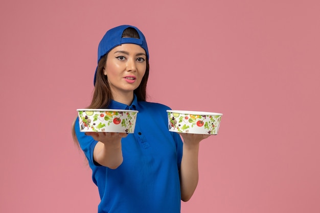 Mensajero femenino de vista frontal en capa uniforme azul sosteniendo cuencos de entrega en la pared rosa claro, servicio de entrega de empleados