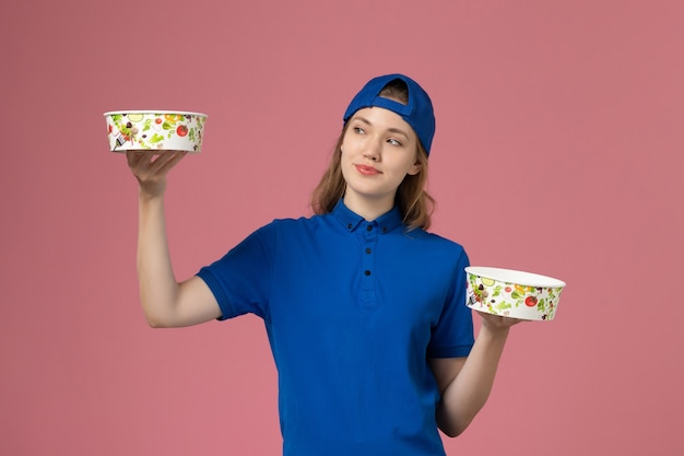 Mensajero femenino de vista frontal en capa uniforme azul sosteniendo cuencos de entrega en la pared rosa, trabajo de niña empleada de entrega de servicio