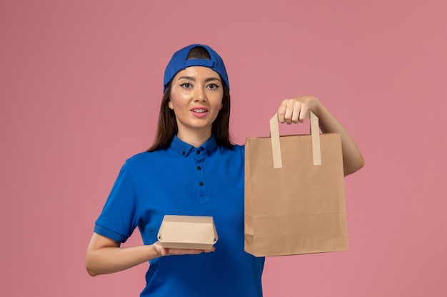 Mensajero femenino de vista frontal en capa uniforme azul sosteniendo diferentes paquetes de entrega en la pared rosa, empleado de servicio entregando