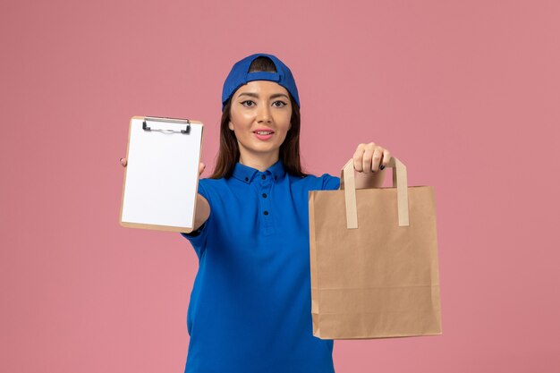 Mensajero femenino de vista frontal en capa uniforme azul sosteniendo el paquete de papel de entrega y el bloc de notas en la pared rosa, trabajador de entrega