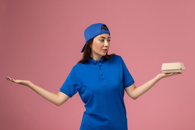 Mensajero femenino de vista frontal con capa uniforme azul sosteniendo un pequeño paquete de entrega en la pared rosa, empresa de niña trabajadora de entrega de servicio de empleado