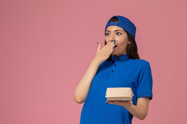 Mensajero femenino de vista frontal con capa uniforme azul sosteniendo un pequeño paquete de entrega en la pared rosa, empresa de niña trabajadora de trabajo de entrega de servicio de empleado