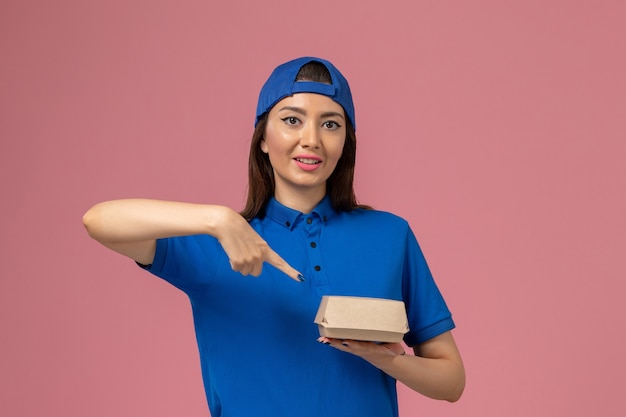 Mensajero femenino de vista frontal con capa uniforme azul sosteniendo un pequeño paquete de entrega en la pared rosa, empresa de trabajo de entrega de servicios para empleados