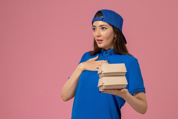Mensajero femenino de vista frontal en capa uniforme azul sosteniendo un pequeño paquete de entrega en la pared rosa, servicio de trabajo de empleado entrega de trabajo