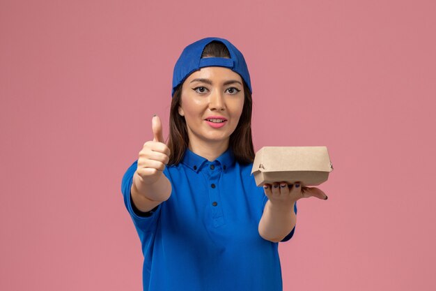 Mensajero femenino de vista frontal en capa uniforme azul sosteniendo un pequeño paquete de entrega en la pared rosa, trabajo de servicio del empleado