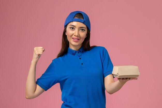 Mensajero femenino de vista frontal en capa uniforme azul sosteniendo un pequeño paquete de entrega en la pared rosa, trabajo de trabajo de entrega de servicio de empleado