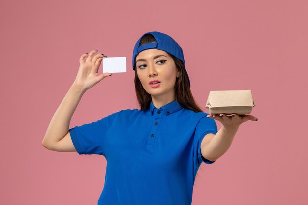 Mensajero femenino de vista frontal con capa uniforme azul sosteniendo un pequeño paquete de entrega con tarjeta de plástico en la pared de color rosa claro, entrega de servicios de trabajo para empleados