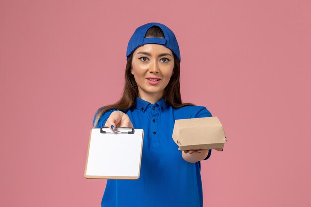 Mensajero femenino de vista frontal con capa uniforme azul sosteniendo un pequeño paquete de entrega vacío con bloc de notas en la pared rosa claro, entrega de servicios de empleados de trabajo