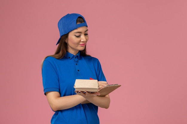 Mensajero femenino de vista frontal con capa uniforme azul sosteniendo un pequeño paquete de entrega vacío con bloc de notas en la pared rosa claro, entrega de trabajo de la empresa de servicios para empleados