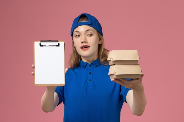 Mensajero femenino de vista frontal en capa uniforme azul sosteniendo pequeños paquetes de comida de entrega y bloc de notas en el empleado de servicio de trabajo de entrega de fondo rosa