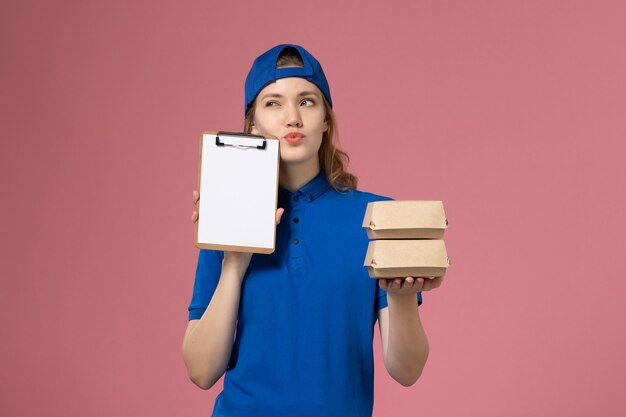 Mensajero femenino de vista frontal con capa uniforme azul sosteniendo pequeños paquetes de comida de entrega y bloc de notas pensando en el empleado del servicio de entrega de fondo rosa