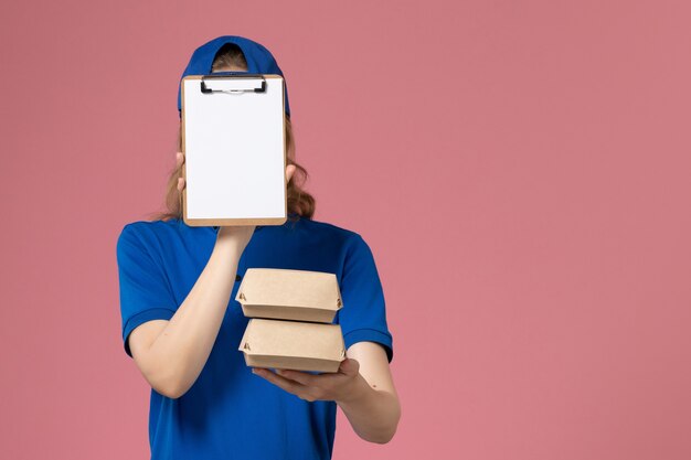 Mensajero femenino de vista frontal con capa uniforme azul sosteniendo pequeños paquetes de comida de entrega y bloc de notas sobre fondo rosa claro empleado del servicio de entrega