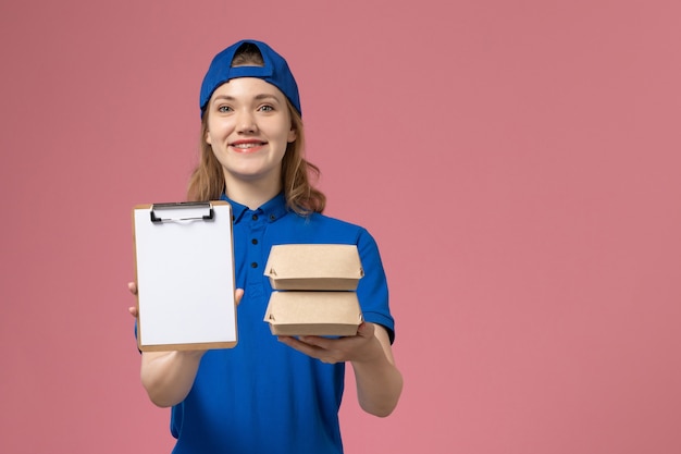 Mensajero femenino de vista frontal en capa uniforme azul sosteniendo pequeños paquetes de comida de entrega y bloc de notas sobre fondo rosa empleado de servicio de entrega