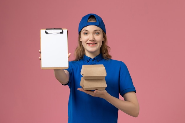 Mensajero femenino de vista frontal en capa uniforme azul sosteniendo pequeños paquetes de comida de entrega y bloc de notas sobre fondo rosa trabajo de niña empleada de servicio de entrega