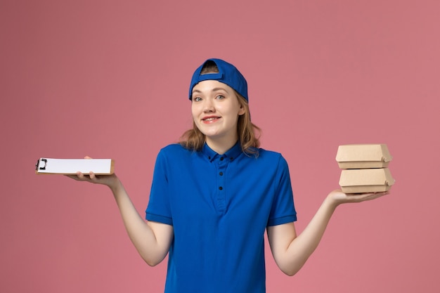 Mensajero femenino de vista frontal en capa uniforme azul sosteniendo pequeños paquetes de comida de entrega y bloc de notas en el trabajo de trabajo de empleado de servicio de entrega de fondo rosa