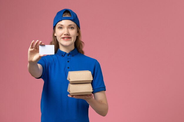 Mensajero femenino de vista frontal en capa uniforme azul sosteniendo pequeños paquetes de comida de entrega y tarjeta sobre fondo rosa trabajo de empleado de entrega de servicio