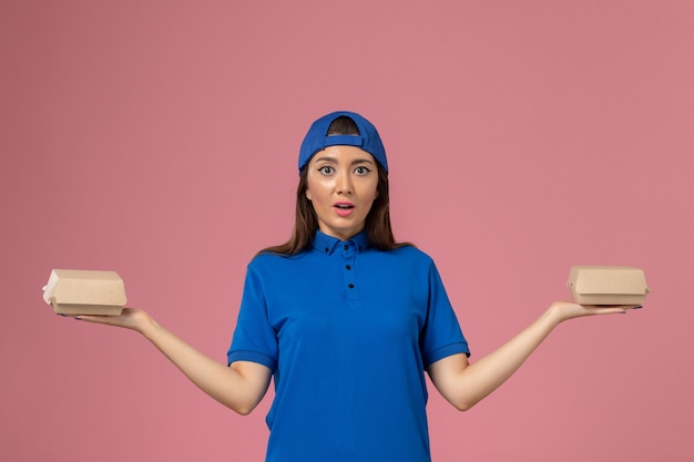 Mensajero femenino de vista frontal con capa uniforme azul sosteniendo pequeños paquetes de entrega en la pared rosa, entrega de servicio de chica empleada